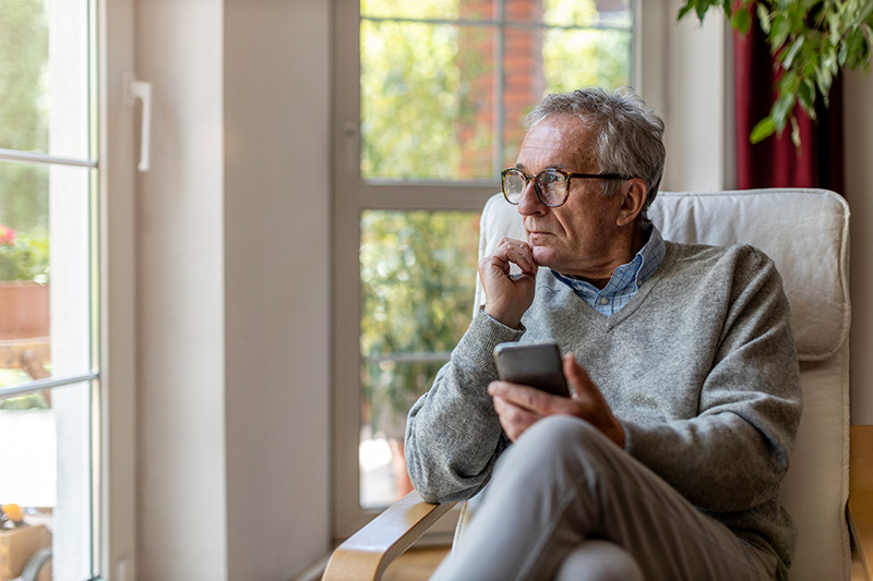 senior; elderly; grandfather; man; old; pensioner; retired; holding; mobile phone; smartphone; technology; window; looking; waiting; stress; loneliness; despair; tired; lonely; alone; sad; unhappy; depression; thinking; thoughtful; trapped; illness; uncertainty; anxiety; worried; looking through window; avoidance; stay at home; using; communication; connected; calling; using phone; retirement; aged; mature; home; house; male; people; lifestyle; domestic life; real people; 