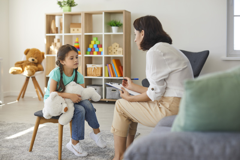 Depressed girl talking to psychologist during therapy session in cozy modern office. Supportive understanding therapist with clipboard listening to child, dealing with behavioral problems