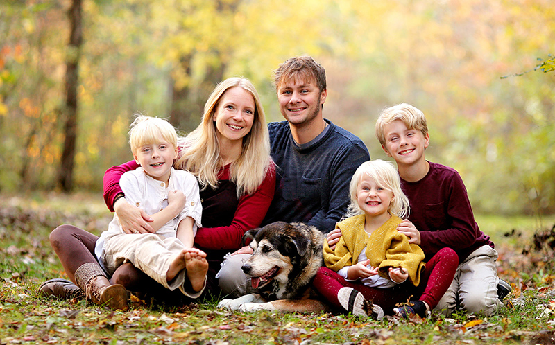 A portrait of a happy young family with three cute, smiling, children and their pet dog sitting outside in the woods on an Autumn day.