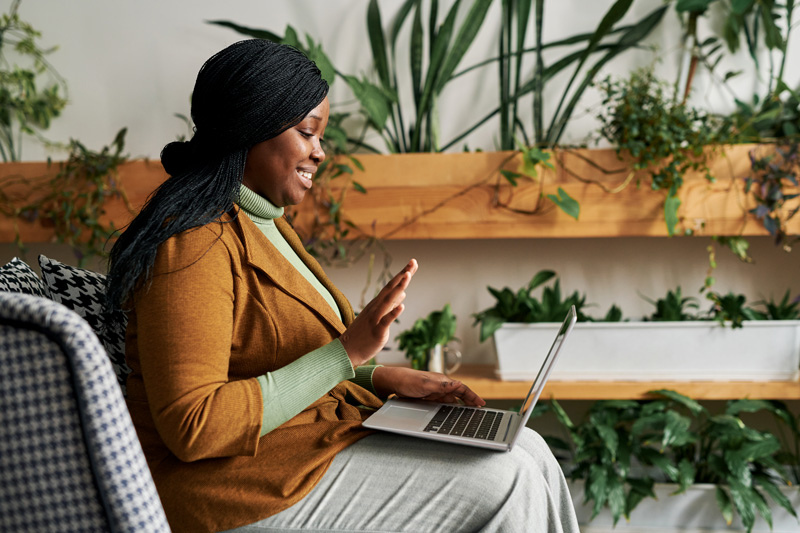 Woman with a laptop on her knees waving hand to her therapist during an online session