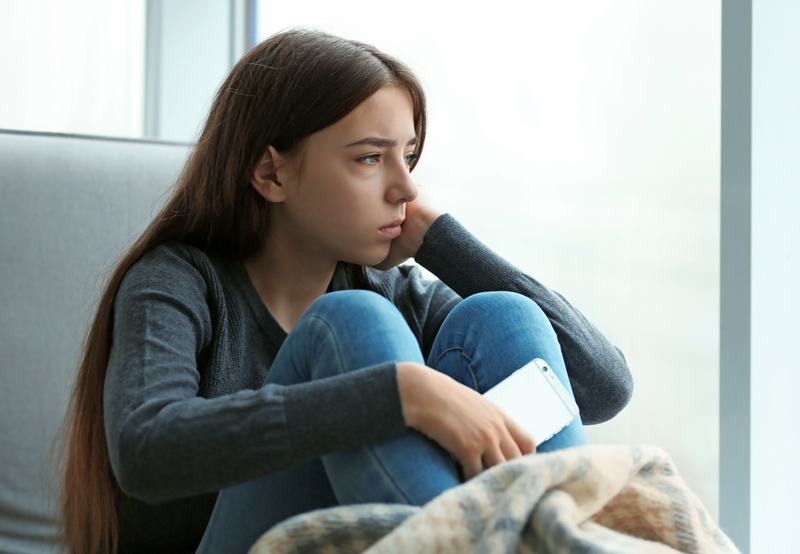 Upset teenage girl with smartphone sitting at window indoors. 