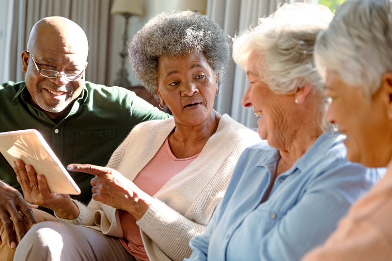 Three happy diverse senior woman and african american male friend sitting on sofa and using tablet