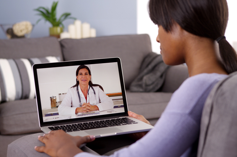 a person sitting on a couch and participating in a virtual medical consultation on a laptop. The screen displays a female doctor smiling, wearing a white coat, with a stethoscope around her neck. The person viewing the screen appears focused on the video call, with the laptop resting on their lap. The background in the room is softly lit, with some home decor, such as a plant and candles, visible in the background. The overall scene suggests a comfortable and modern setting for a telemedicine appointment.