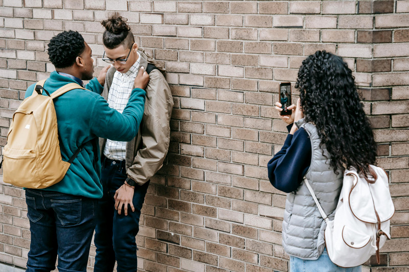 One boy holding another against the wall while a girl films on her phone