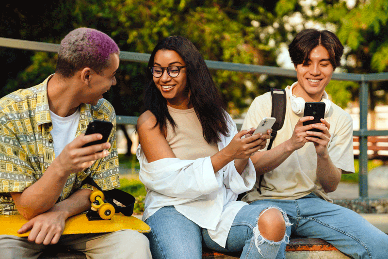 Three happy teens on their phones sitting together outside