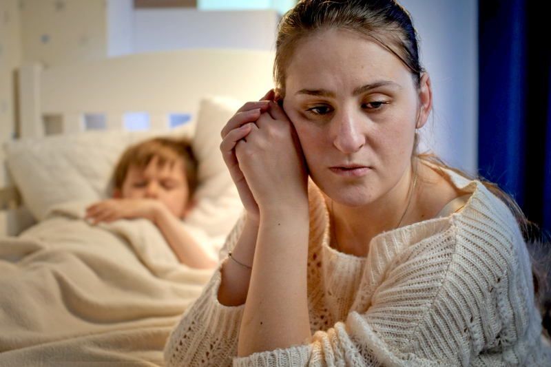 Upset and stressed mother crying next to bed of her little son sleeping at night. 